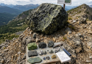 Photographie de paysage prise sur un affleurement rocheux surplombant une vaste vallée et des montagnes lointaines. Plusieurs gros échantillons de roches, notamment des dalles vert jade et des blocs tachetés, sont regroupés au premier plan avec un marteau de géologue posé à côté.